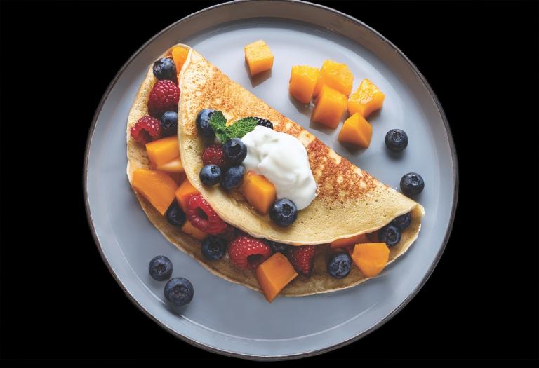 A pancake wrapped around cantaloupe chunks, raspberries, and blueberries, with a dollop of yogurt on top.